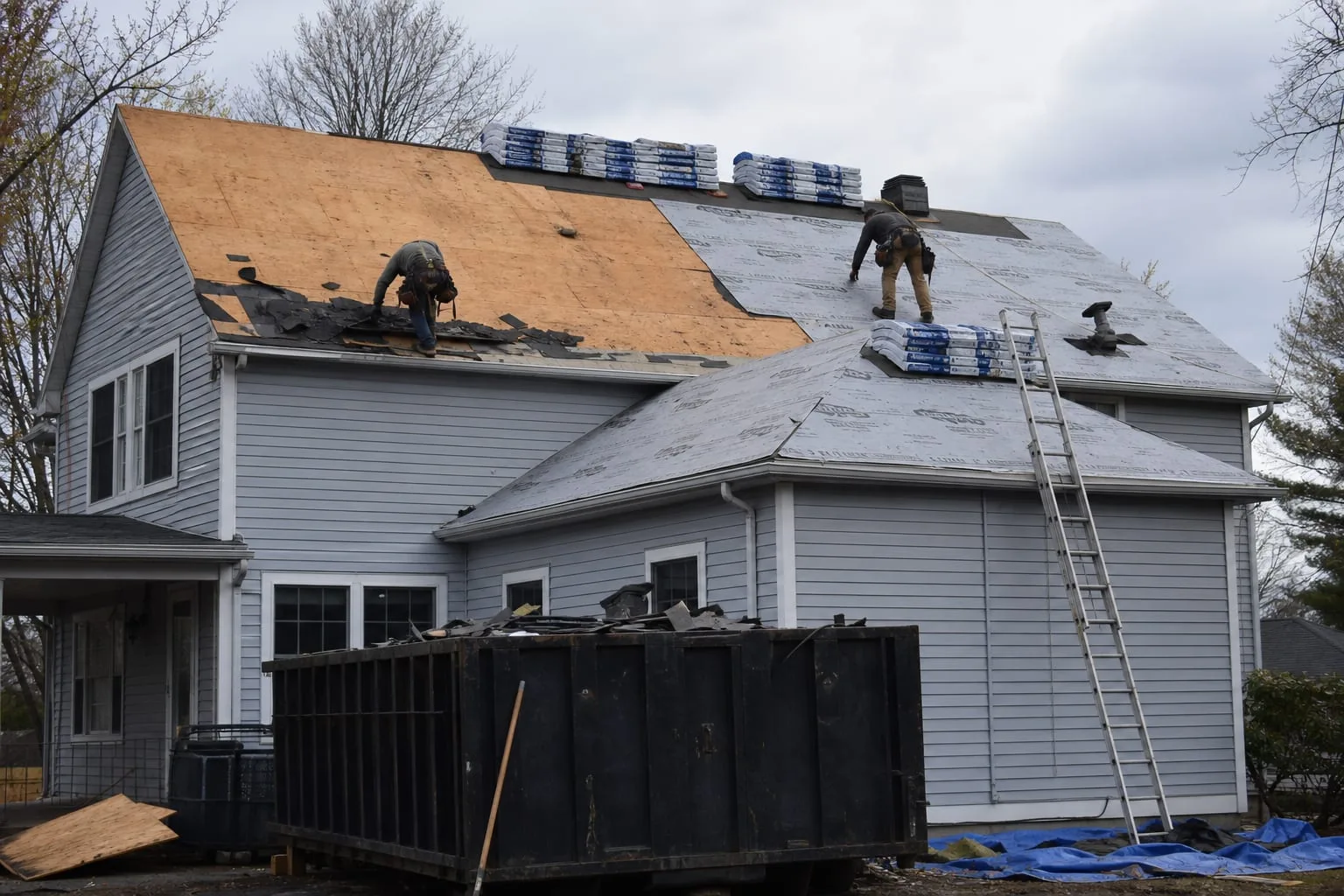A roof tear-off in progress on a two-story house, showing exposed decking on one section, new underlayment on another, and stacks of bundled shingles on the...
