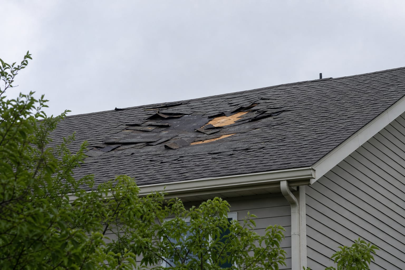 A damaged section of shingle roof photographed from ground level against an overcast sky