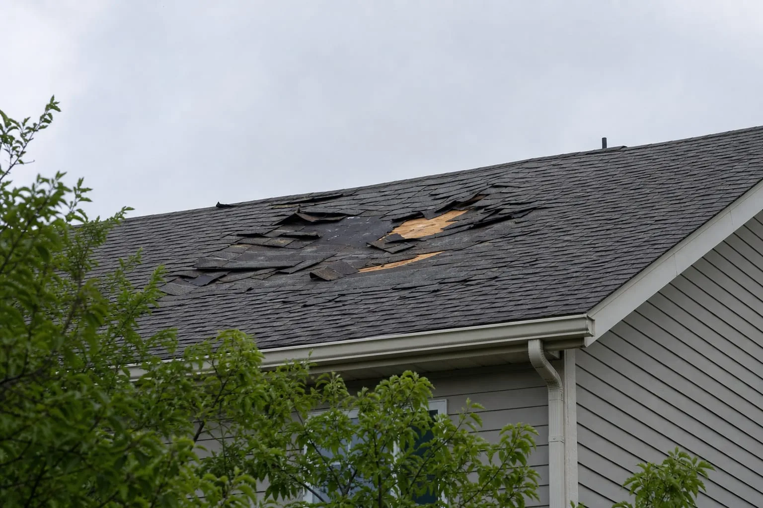 A damaged section of shingle roof photographed from ground level against an overcast sky